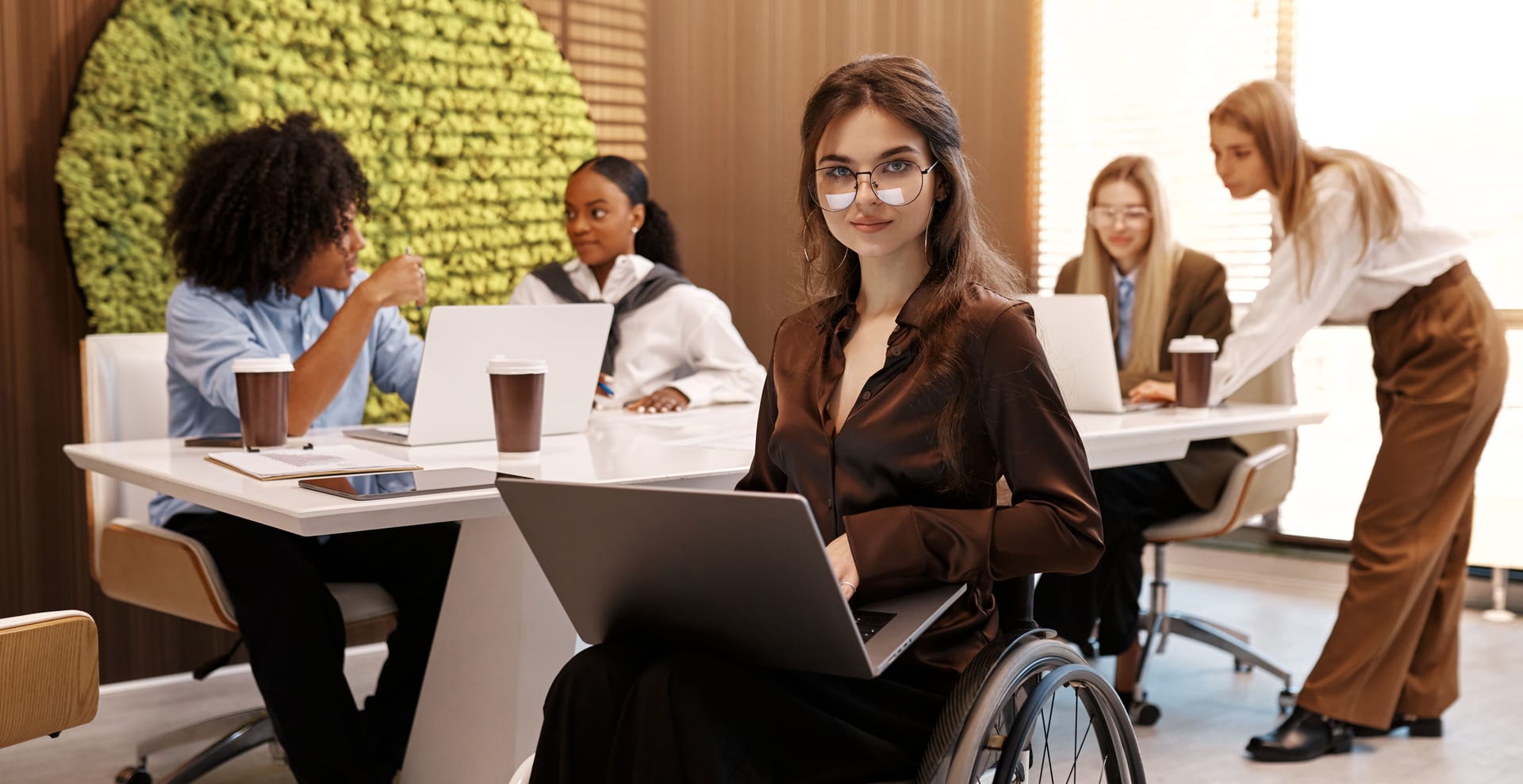 Mitarbeiterin im Rollstuhl arbeitet mit Laptop in einem inklusiven Team in einem modernen Büro.