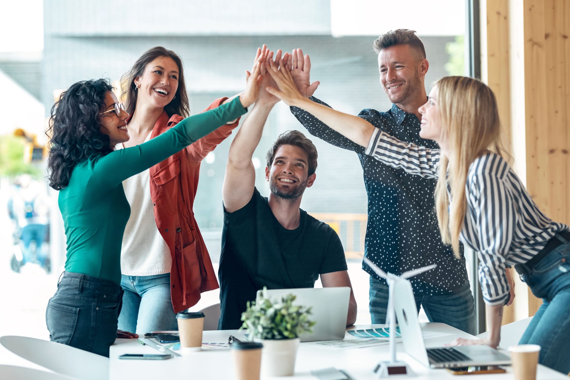 Fünf junge Berufstätige im Büro geben sich ein gemeinsames High-Five über einem Tisch mit Laptops und Kaffee und wirken motiviert und erfolgreich.
